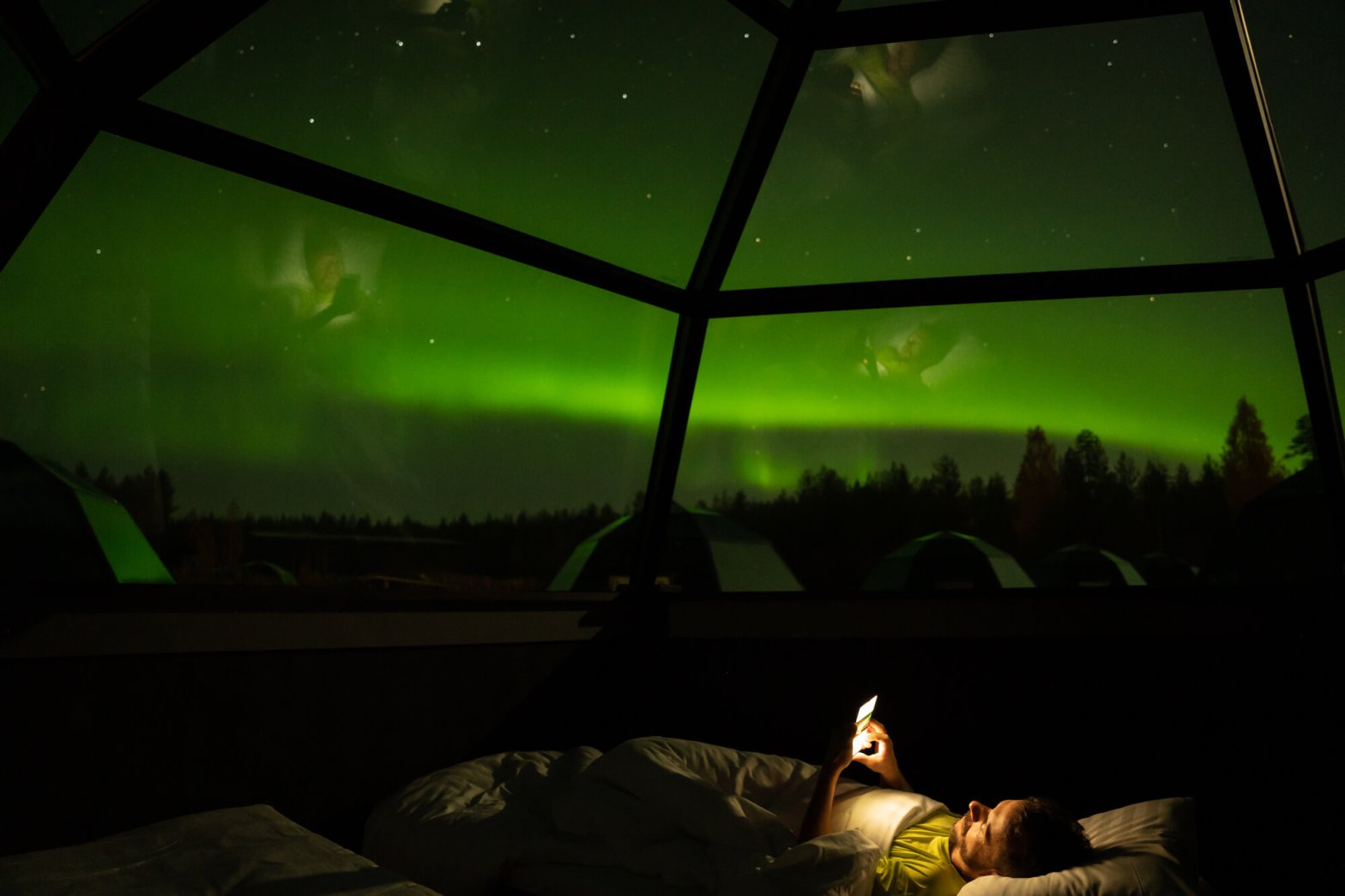 Person lying down in a glass igloo under northern lights